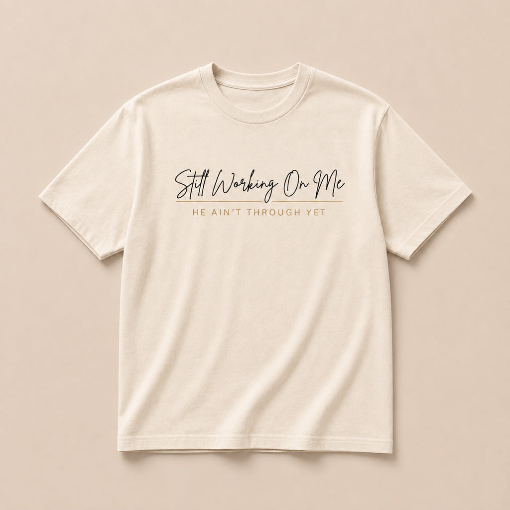 Still Working On Me cream t-shirt laid flat, script lettering with He Ain't Through Yet beneath