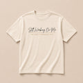 Still Working On Me cream t-shirt laid flat, script lettering with He Ain't Through Yet beneath
