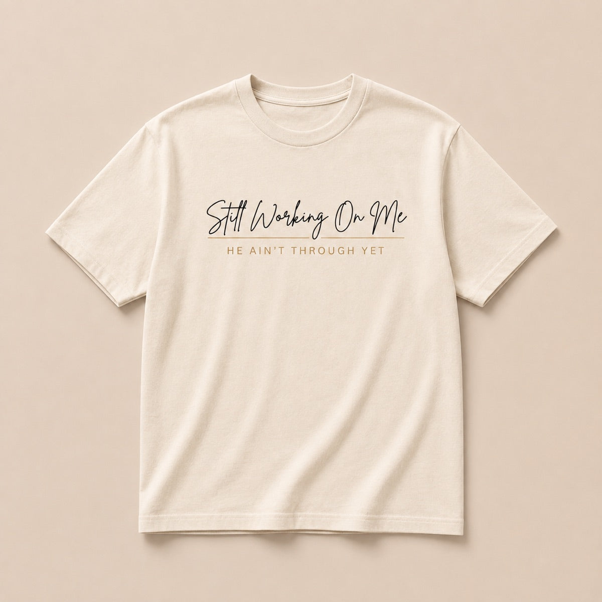 Still Working On Me cream t-shirt laid flat, script lettering with He Ain't Through Yet beneath