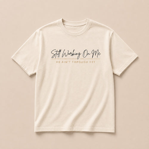 Still Working On Me cream t-shirt laid flat, script lettering with He Ain't Through Yet beneath